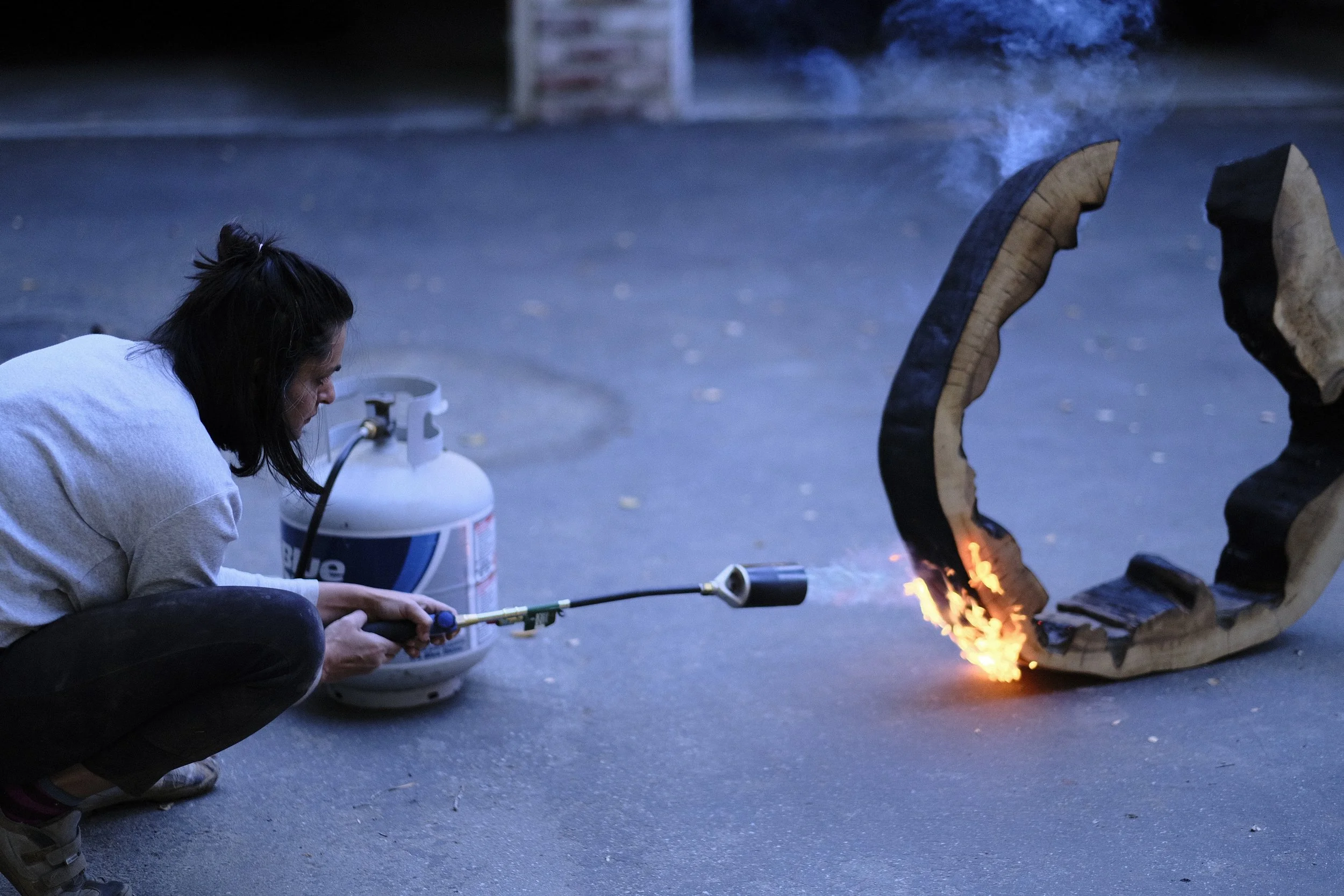The artist crouched with a propane torch, flame against the base of a curved wood form — charring in progress.