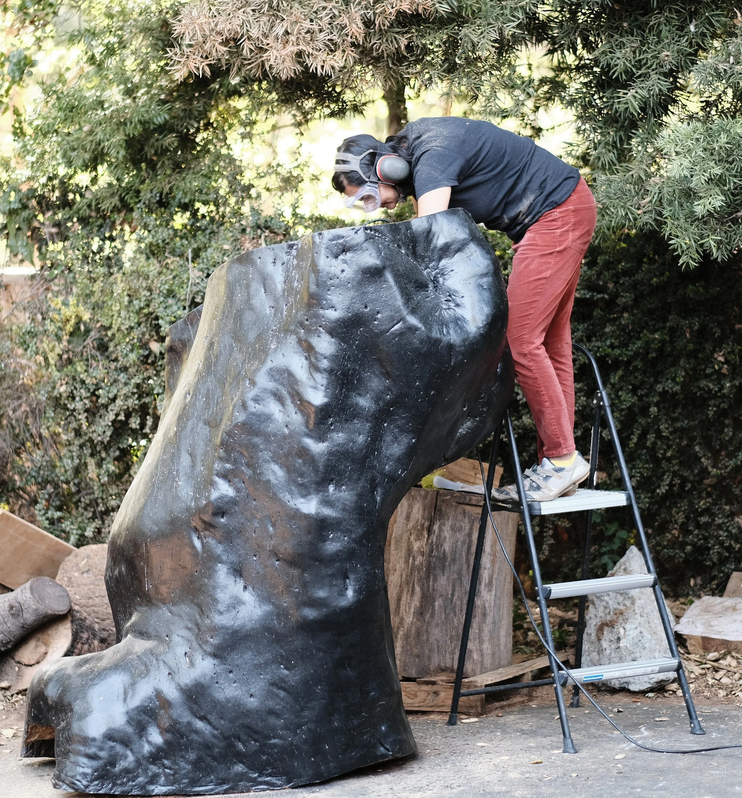 The artist on a ladder, carving the top of a large blackened wood form outdoors.