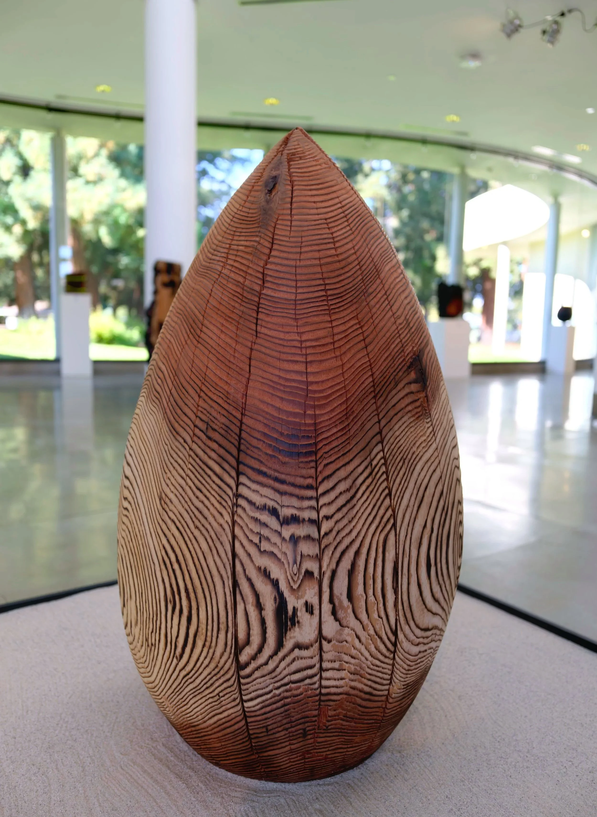Monsoon — a carved redwood teardrop resting in a bed of sand, in a bright gallery.