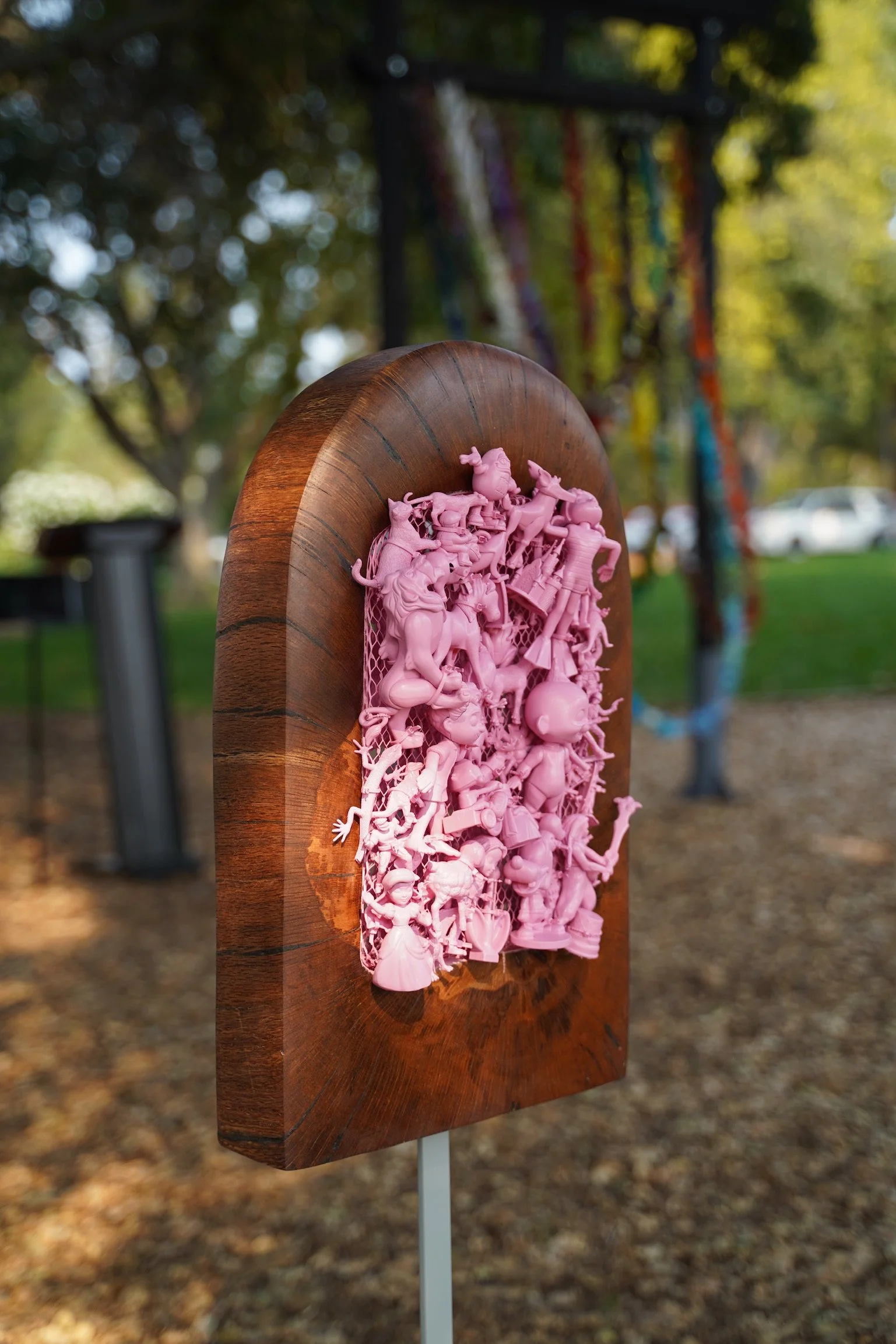 What’s At Play — an arched wood panel holding a dense relief of pink spray-painted plastic toys, outdoors.