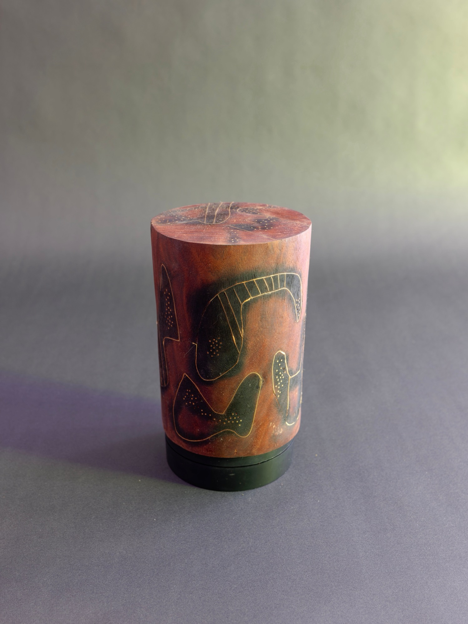Dream — a carved cylindrical wood sculpture, its rust-red surface inscribed with dark biomorphic figures, on a black base against a grey-green studio backdrop.